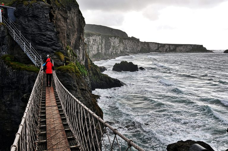 Visitors crossing Carrick-a-Rede rope bridge above rough seas, with steep coastal cliffs beneath an overcast sky in County Antrim, Northern Ireland.