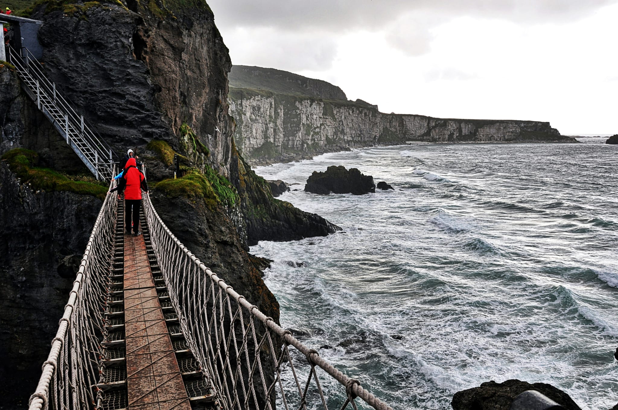 Visitors crossing Carrick-a-Rede rope bridge above rough seas, with steep coastal cliffs beneath an overcast sky in County Antrim, Northern Ireland.